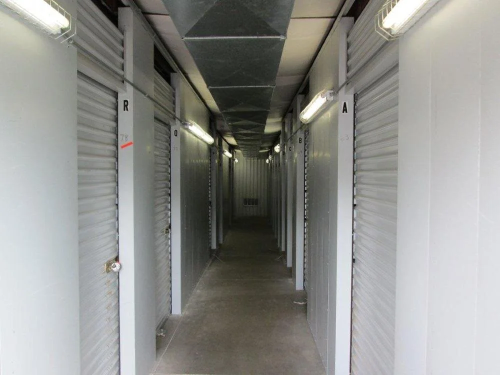 An indoor storage unit hallway at All Hours Storage with metal roll up doors on both sides and climate control ducting overhead