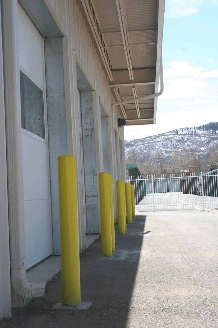Roll-up storage unit doors along the parking apron at All Hours Storage, protected by yellow bollards